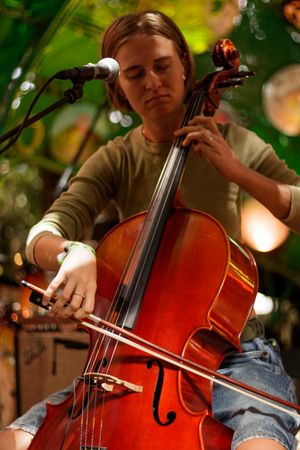 Young musician playing cello on stage with microphone, performing live at an outdoor music event.