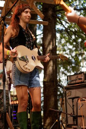 Woman playing white electric guitar on outdoor stage, wearing denim shorts and green boots at a music festival.