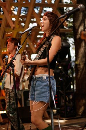 Young woman singing and playing guitar on outdoor stage in denim shorts and black crop top.