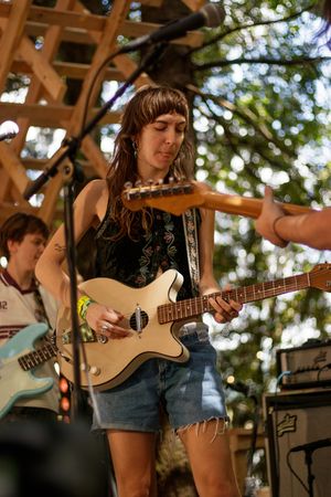Woman playing white electric guitar on outdoor stage in denim shorts and black top, with band member visible behind her.