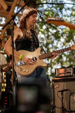 Young woman playing a cream electric guitar on an outdoor stage surrounded by green trees at a music festival.