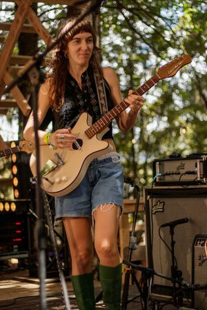 Female guitarist performing outdoors in denim shorts and green socks, playing a cream electric guitar on stage.