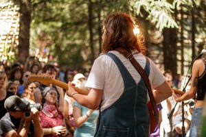 Guitarist in overalls performing outdoors with crowd of festival attendees in forest setting.