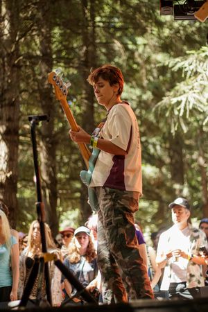 Young guitarist performing on outdoor stage in camouflage pants, playing teal electric guitar with forest backdrop.