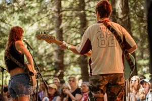 Two musicians perform on outdoor stage at forest festival, guitarist in number 82 jersey with crowd.