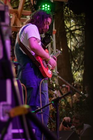 Musician with long hair plays red electric guitar on outdoor stage, wearing overalls and white shirt under green stage lights.