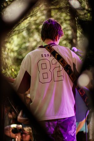 Guitarist on outdoor stage wearing white ESPA 82 jersey with colorful guitar strap, performing among trees with purple stage lighting.