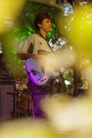 Young guitarist performing outdoors on stage, holding electric guitar under green and purple stage lights filtered through leaves.