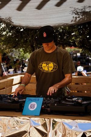DJ Spmitch wearing Wu-Tang shirt spins vinyl records on turntables at an outdoor music event under a shaded canopy.