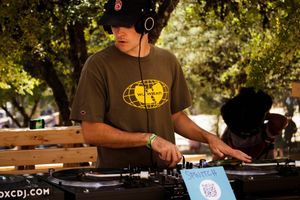A DJ wearing headphones and a bucket hat mixes tracks on turntables at an outdoor music event.