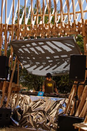 DJ performing under a wooden lattice structure outdoors, with speakers and equipment surrounded by crinkled foil decorations.