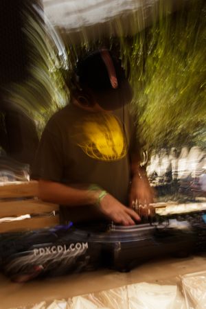 A DJ wearing headphones and a yellow graphic tee spins records outdoors with motion blur effect.
