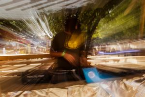 Blurred motion photo of a person sitting under a tree at a vibrant outdoor market at night.