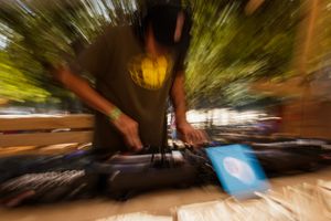 DJ mixing music outdoors with motion blur effect, wearing headphones at a turntable setup in a sunny setting.