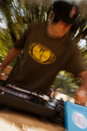 A DJ wearing headphones and a dark t-shirt with a yellow globe logo spins records outdoors among trees.