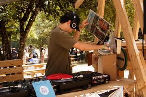 Outdoor DJ spinning vinyl records on turntables at a sunny park event, wearing headphones and holding an album.