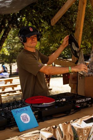 DJ spinning vinyl records outdoors at a festival, wearing headphones and a cap, with turntables and PDXCDJ signage visible.