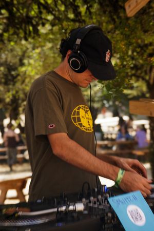 A DJ wearing headphones and a cap mixing records outdoors at a sunny park event with trees in the background.
