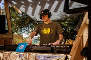 DJ wearing headphones and Wu-Tang shirt mixing vinyl records outdoors at PDXCDJ.com event on sunny day.
