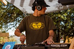 DJ wearing headphones and olive green shirt mixing at outdoor turntable setup with PDXCDJ.COM sign visible.