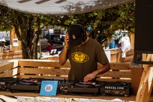 DJ in black cap and olive shirt performing at outdoor turntable event with PDXCDJ.COM branding on display.