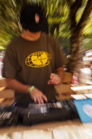 DJ wearing Wu-Tang Clan shirt and headphones mixing music outdoors at a festival with motion blur effect.