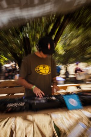 A DJ performs on a keyboard at an outdoor festival, wearing a Wu-Tang Clan shirt, with a zoom blur effect.
