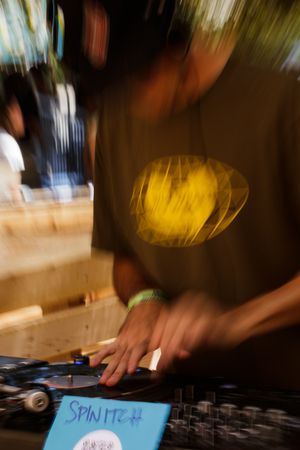 A DJ spinning records at a turntable setup outdoors, wearing a green shirt with a yellow logo, motion blur effect.