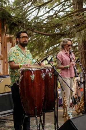 Two musicians perform outdoors, one playing conga drums and another playing clarinet, with saxophones visible on stage.
