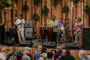 Live band performing outdoors on a wooden stage with congas, guitar, and drums for an engaged crowd.