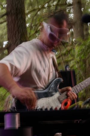 Blurred motion photo of a guitarist playing electric guitar outdoors among trees at a live music performance.