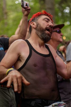 Bearded man with red hair dancing in mesh tank top at outdoor festival.