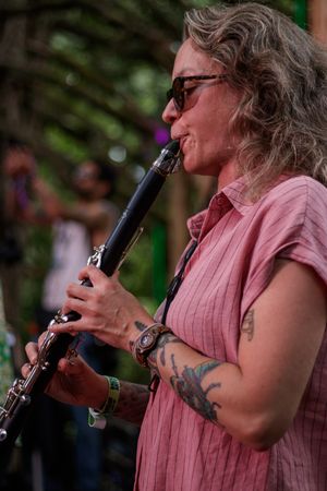 Woman with tattoos playing clarinet outdoors, wearing sunglasses and pink shirt at a music festival.