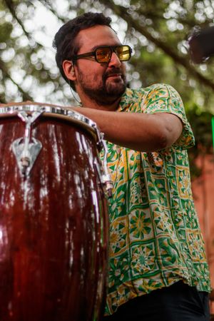 Bearded man in floral shirt and sunglasses playing a conga drum outdoors.