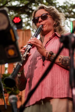 Tattooed woman playing clarinet on stage wearing sunglasses and pink shirt at outdoor music festival.