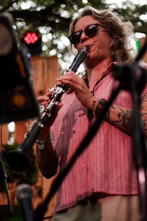 Tattooed woman playing clarinet outdoors, wearing sunglasses and pink shirt at live music performance.