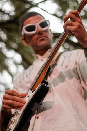 Man in white sunglasses playing electric guitar outdoors, shot from below with bokeh tree background.