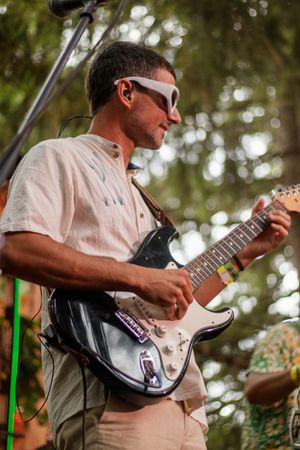 Man in white shirt and sunglasses playing electric guitar on outdoor stage with microphone stand nearby.