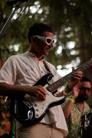 Man in white shirt and stylish white sunglasses plays black electric guitar at outdoor music festival.