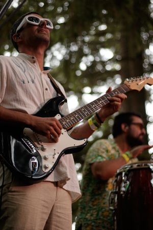 Musician in white sunglasses plays electric guitar outdoors while percussionist plays conga drums in background.