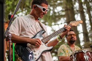 Guitarist in white sunglasses plays Stratocaster on outdoor stage with percussionist in floral shirt behind him.