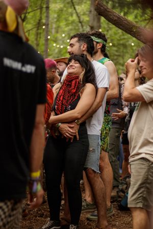 A couple embraces at an outdoor forest festival, surrounded by festivalgoers with string lights in the trees.