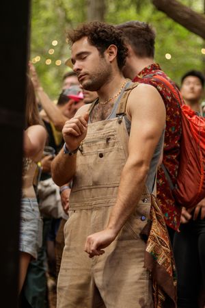 Young man in tan overalls dancing outdoors at a forest festival surrounded by a crowd.