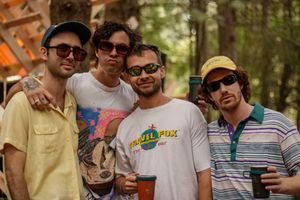 Four friends smiling outdoors at a festival in a wooded area, wearing sunglasses and casual summer clothing.