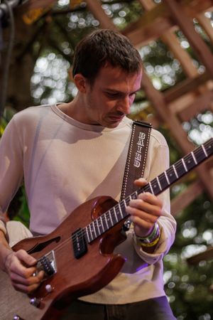 Young man in white sweater playing brown electric guitar outdoors at a music festival.