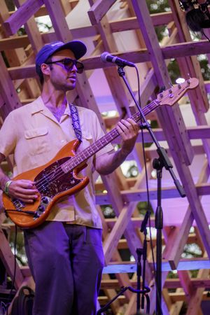 Musician playing orange bass guitar on stage with wooden lattice backdrop under purple lights.