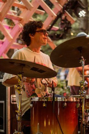 Smiling drummer with curly hair and glasses plays cymbals and drums on an outdoor stage with pink wooden decor.