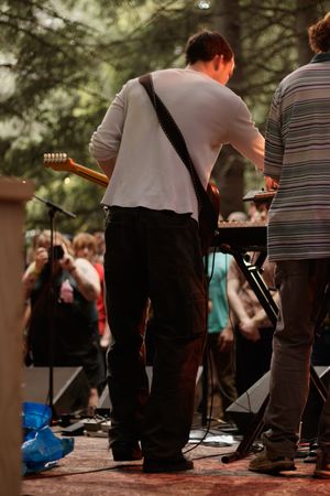 Guitarist performing on outdoor stage viewed from behind with an audience gathered among trees.
