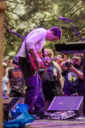Guitarist performing on outdoor stage at music festival, bending over electric guitar with crowd watching behind him.