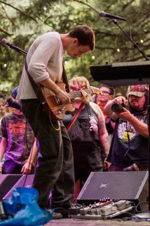Guitarist performs on outdoor stage with crowd watching, using effects pedals, at a forest music festival.
